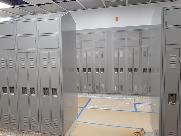 Two-tier standard metal lockers with additional storage above in a locker room, with a gray finish.